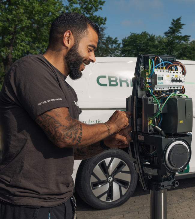 Elektriker udfører service på ladestander med sin elbil i baggrunden