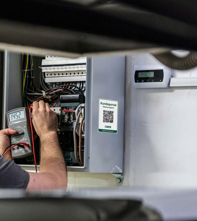 Tekniker udfører lovpligtigt eftersyn på tavle, hvor dokumentationen efterfølgende er tilgængelig i CBREs kundeportal via QR-koden